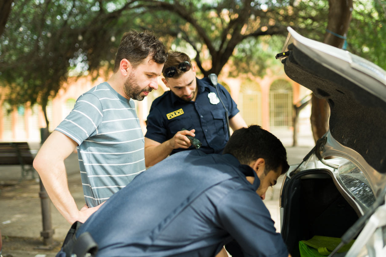 A man stands by as two officers search his vehicle.